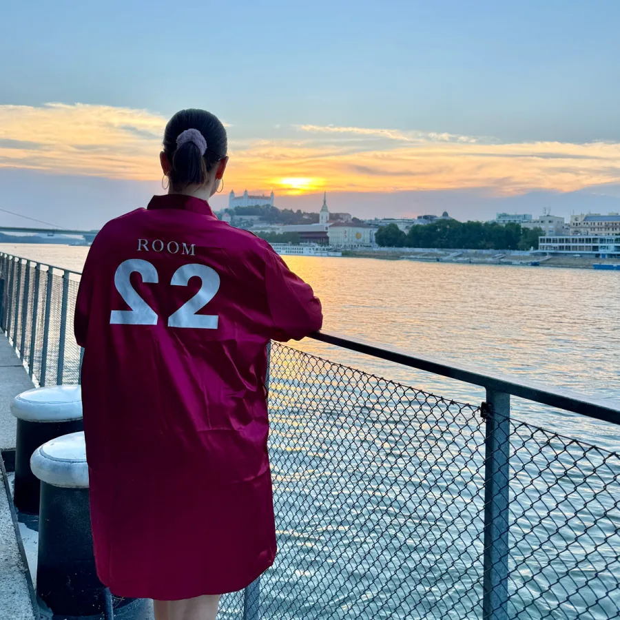Wine-red satin robe with the ROOM 22 sign across the back, worn at sunset by the Danube with Bratislava Castle behind